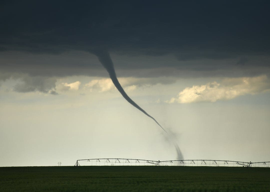 Towering tornado funnel touching down on a green field next to a long metal irrigation pipe under dark clouds.