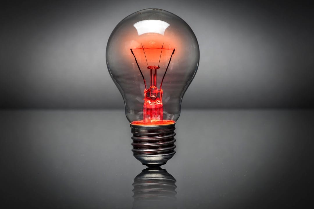 Clear incandescent bulb with a glowing red filament rests on a reflective dark surface against a gray background.