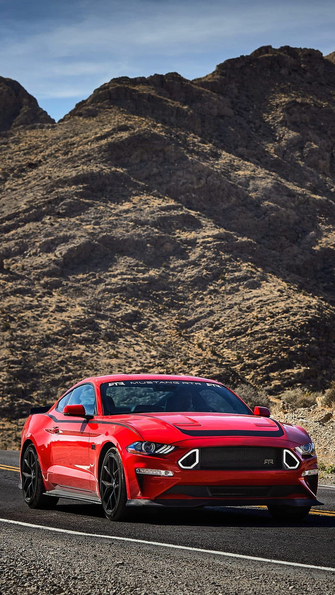 Red Ford Mustang RTR muscle car driving on a desert road between rugged mountains under a clear blue sky.