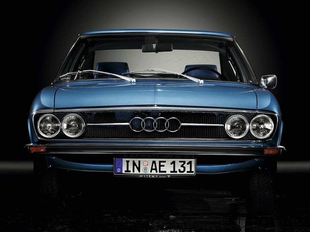 Front view of a vintage blue Audi 100 Coupe S with quad headlights and chrome grille against a dark background.