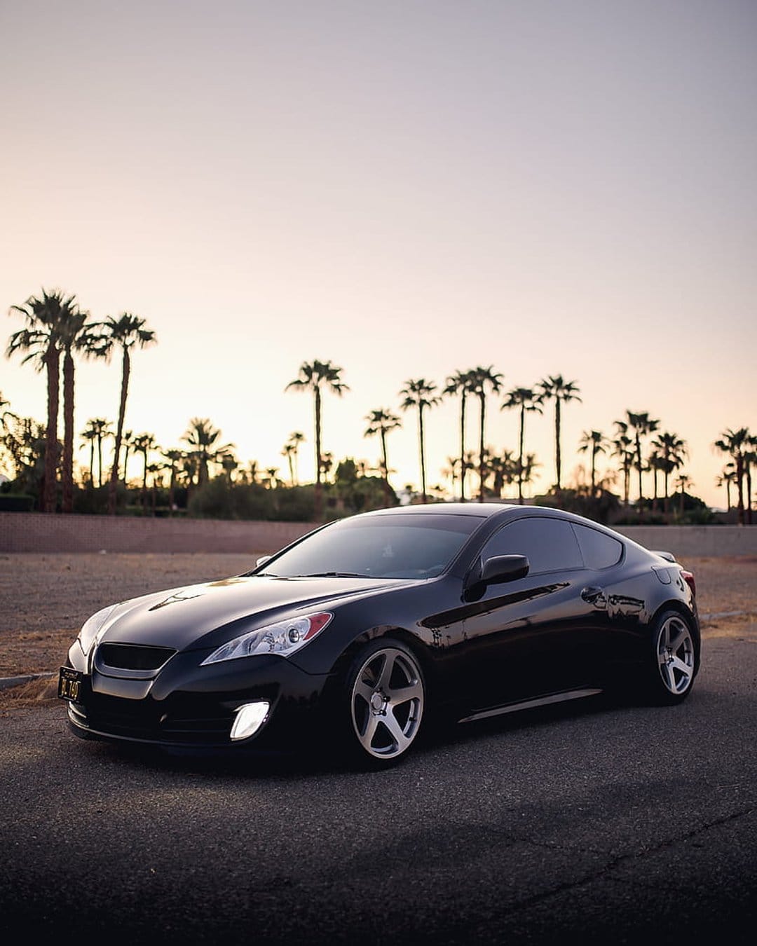 Black Hyundai Genesis Coupe parked on asphalt with palm tree silhouettes against a glowing orange sunset sky.