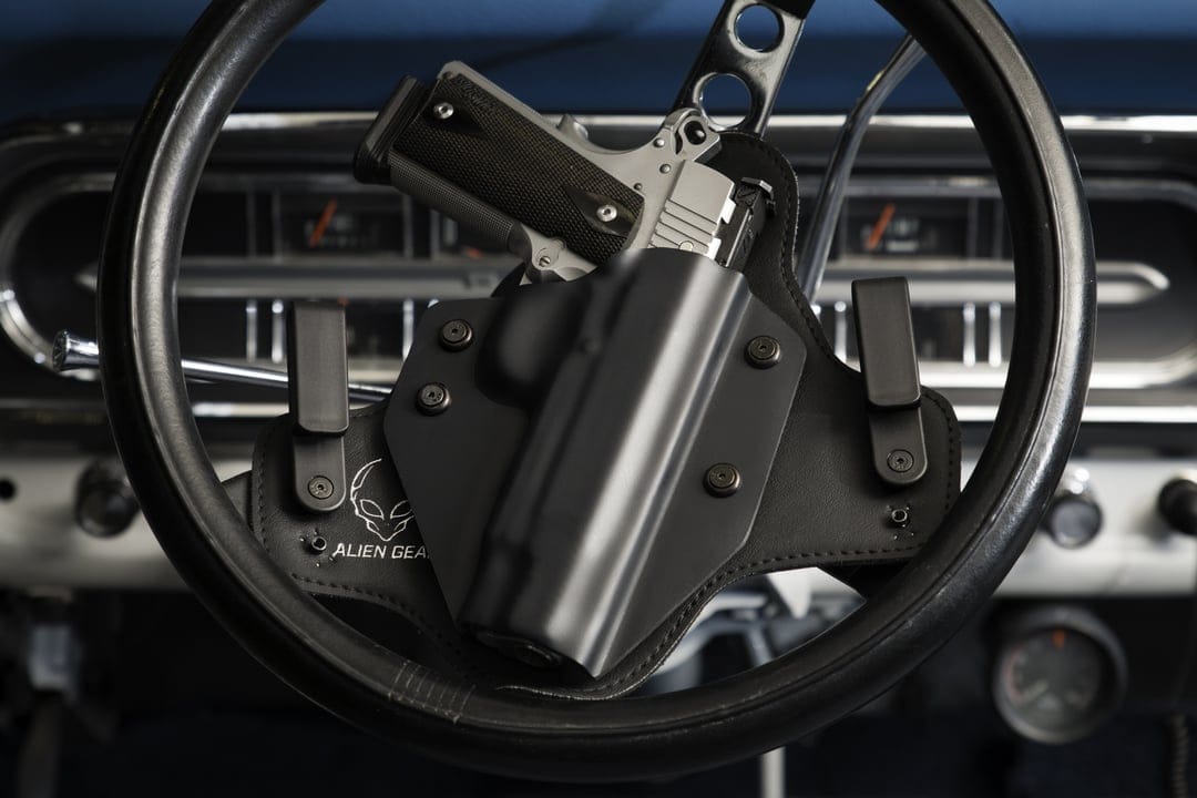 Silver semi-automatic pistol in a black holster mounted to a vintage car steering wheel with leather trim.