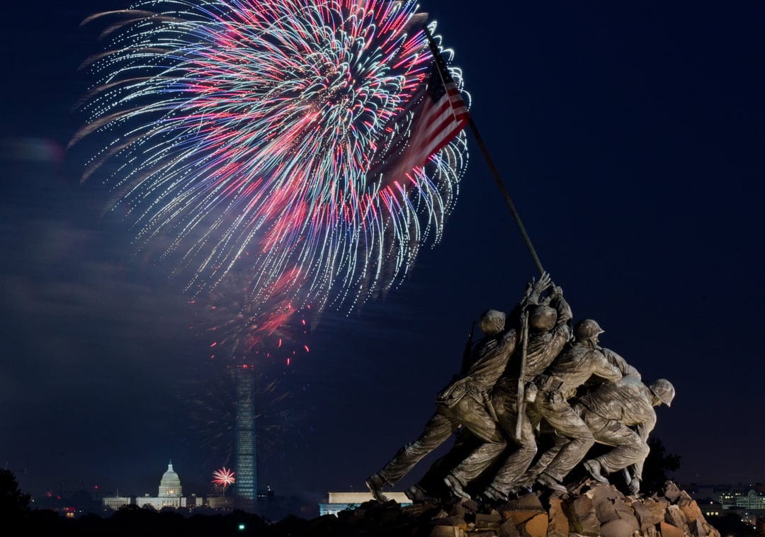 Iwo Jima Memorial statue silhouetted against red and blue fireworks over the Washington D.C. nighttime skyline.