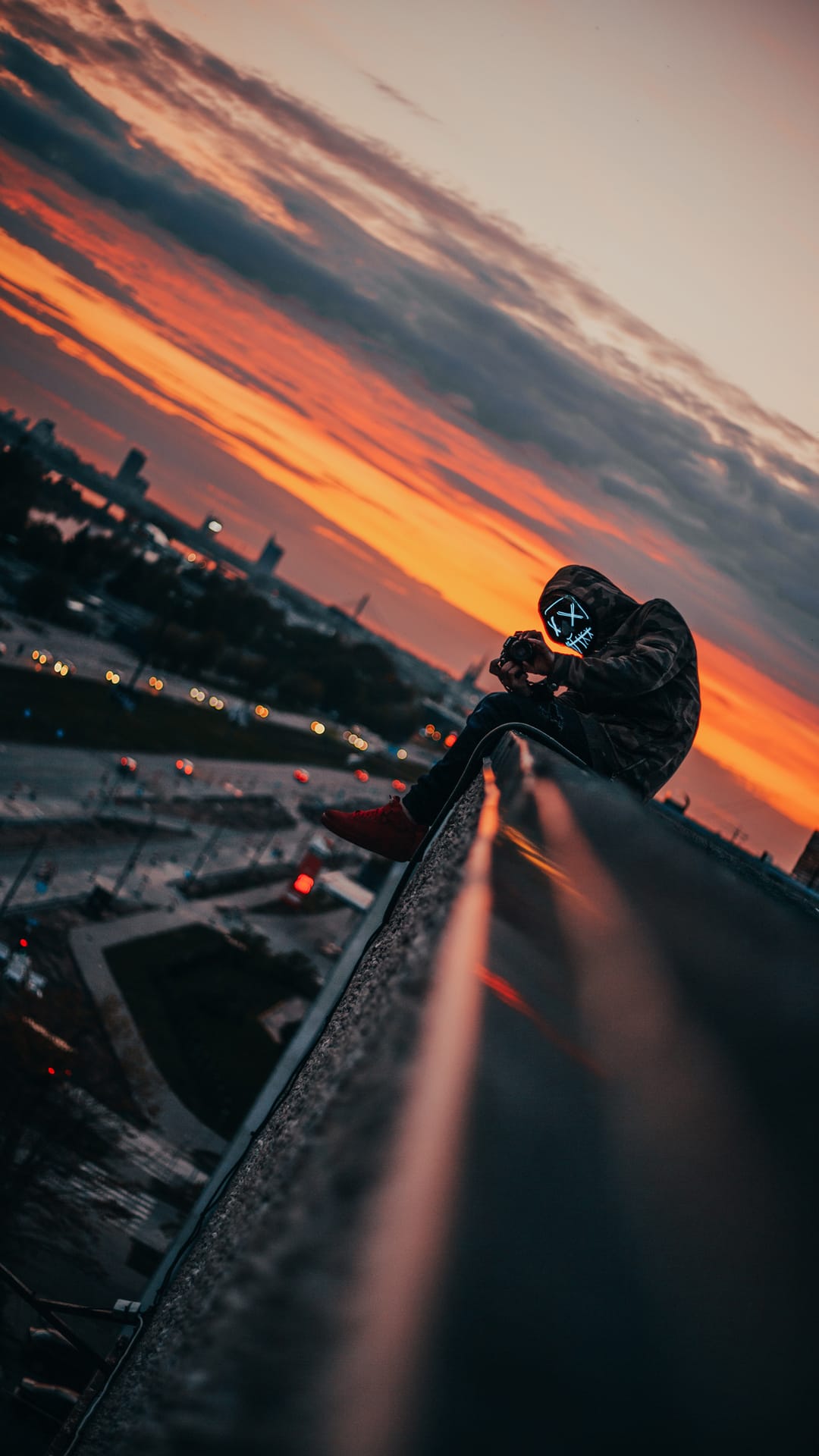 A person wearing a glowing blue X mask sits on a skyscraper ledge overlooking a fiery orange city sunset.