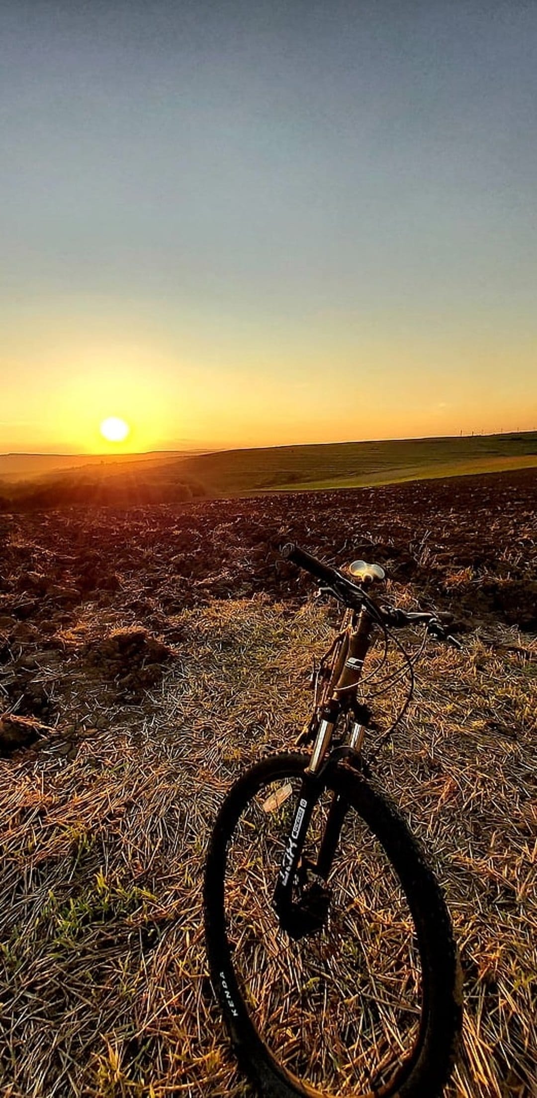 Mountain bike parked in a plowed agricultural field during a golden hour sunset with a soft blue and yellow sky.