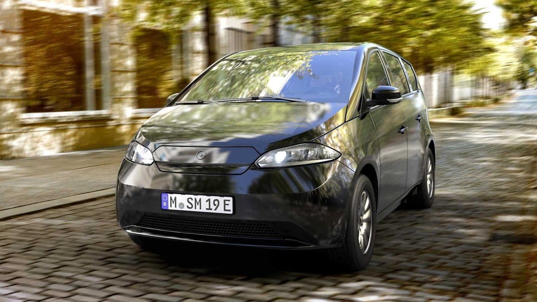 Black Sono Sion solar electric car driving on a cobblestone street with motion-blurred trees and golden sunlight.