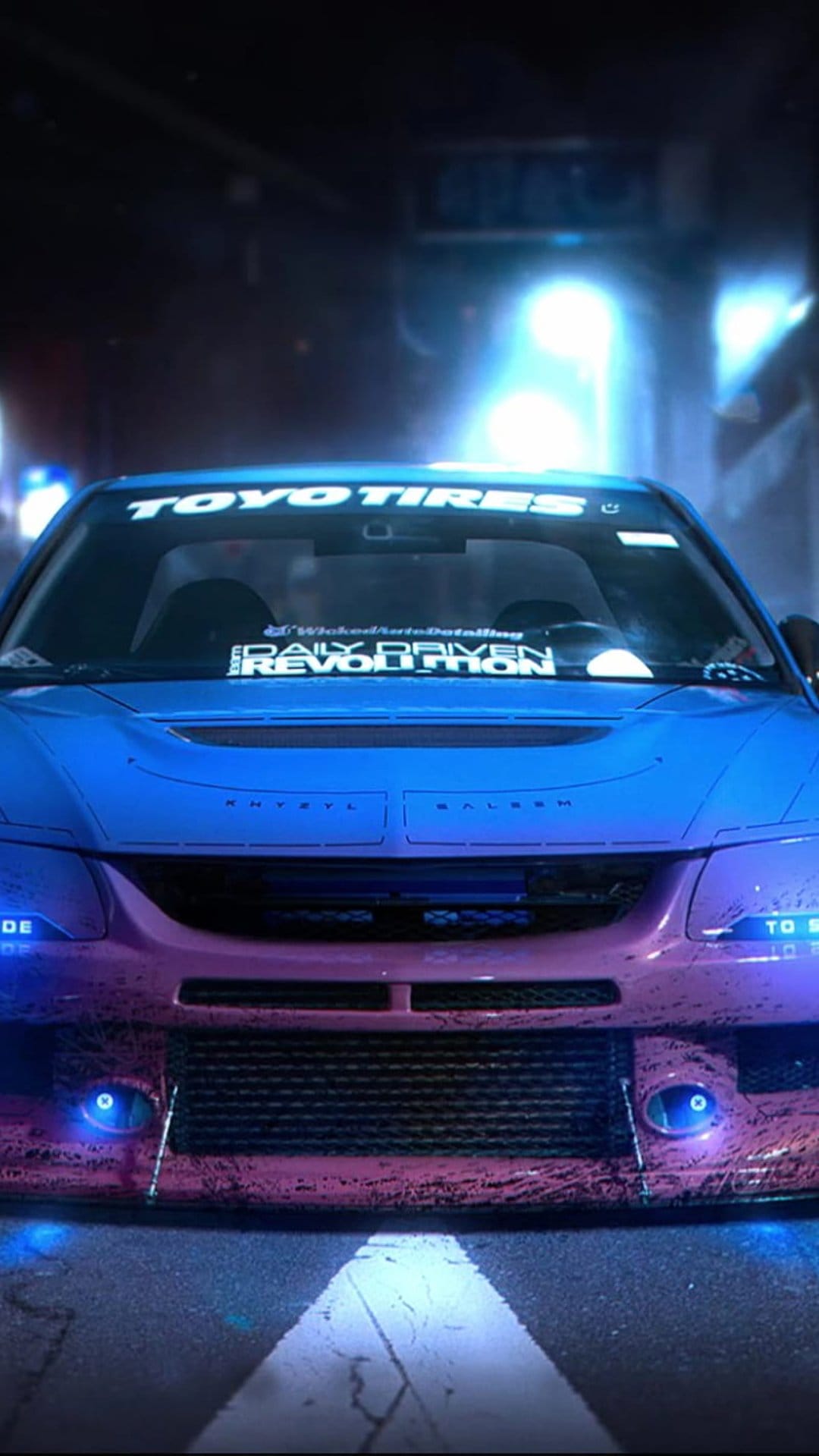 Modified tuner car with blue and pink matte paint and glowing headlights parked on a dark city street at night.