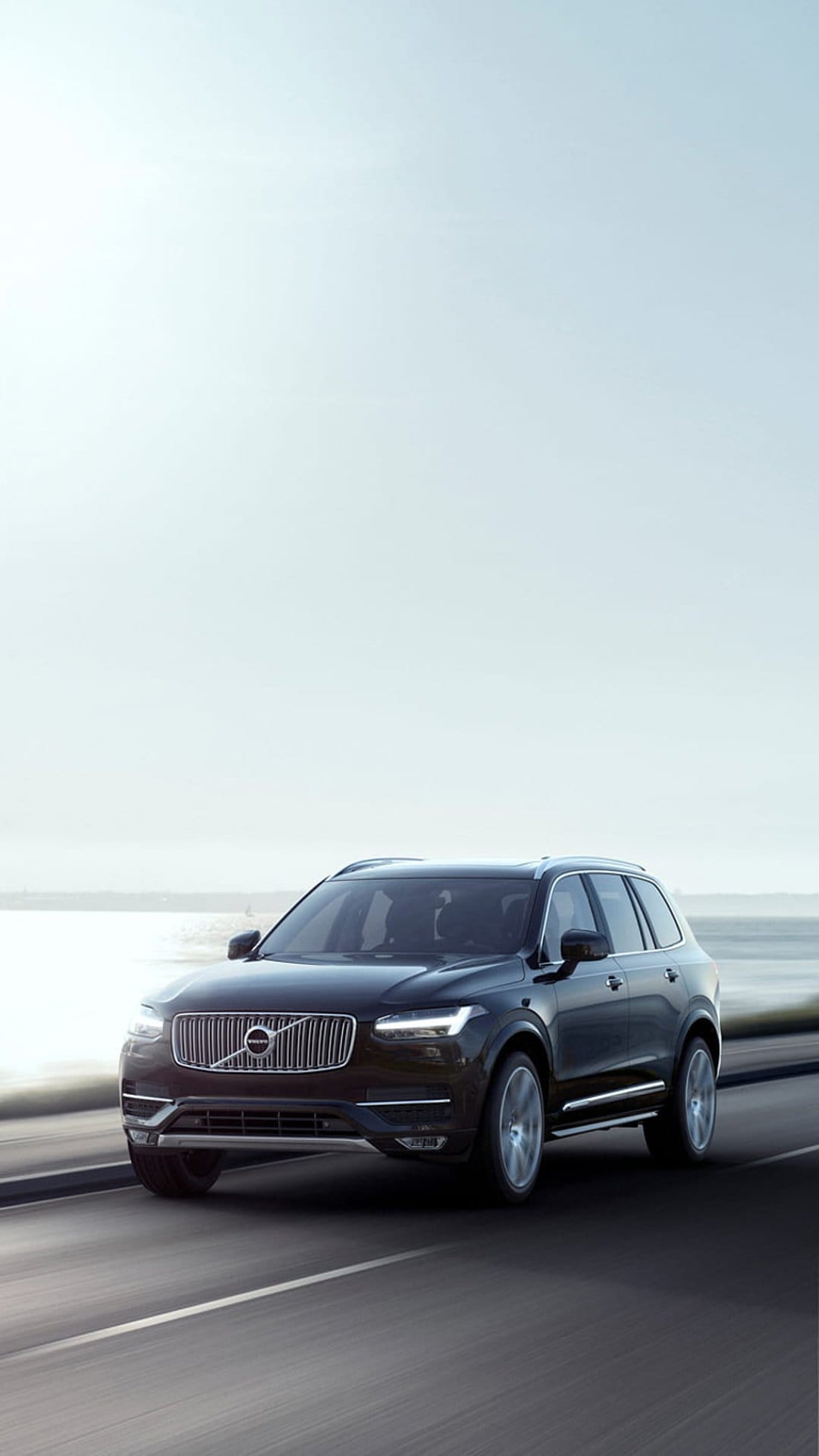 Dark Volvo XC90 SUV drives on a winding coastal road with glowing headlights under a hazy, overcast seaside sky.