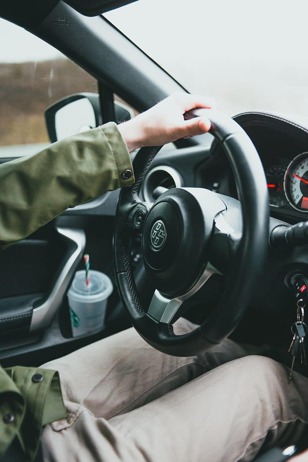 Hands grip a car steering wheel next to a coffee cup in a door pocket, viewed from the driver's perspective.