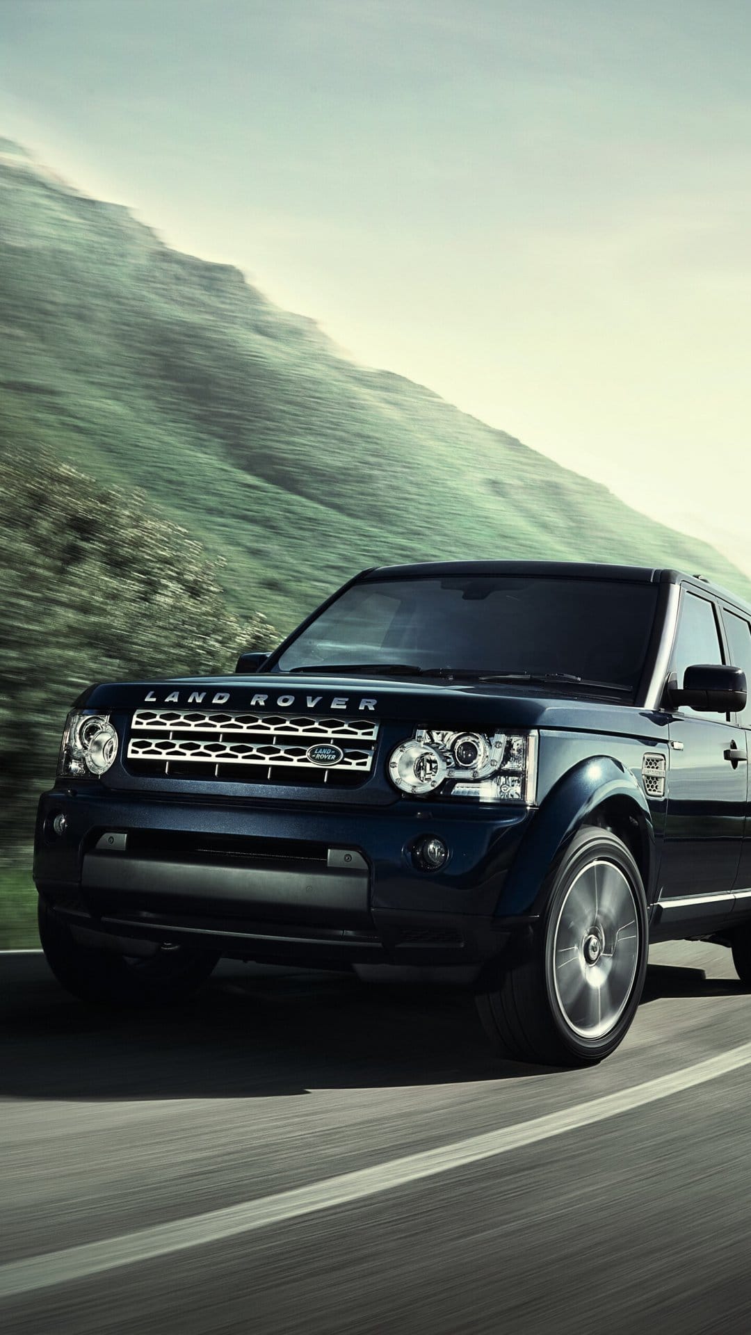 Dark blue Land Rover SUV driving on a mountain road with motion blur and green hills in the background.