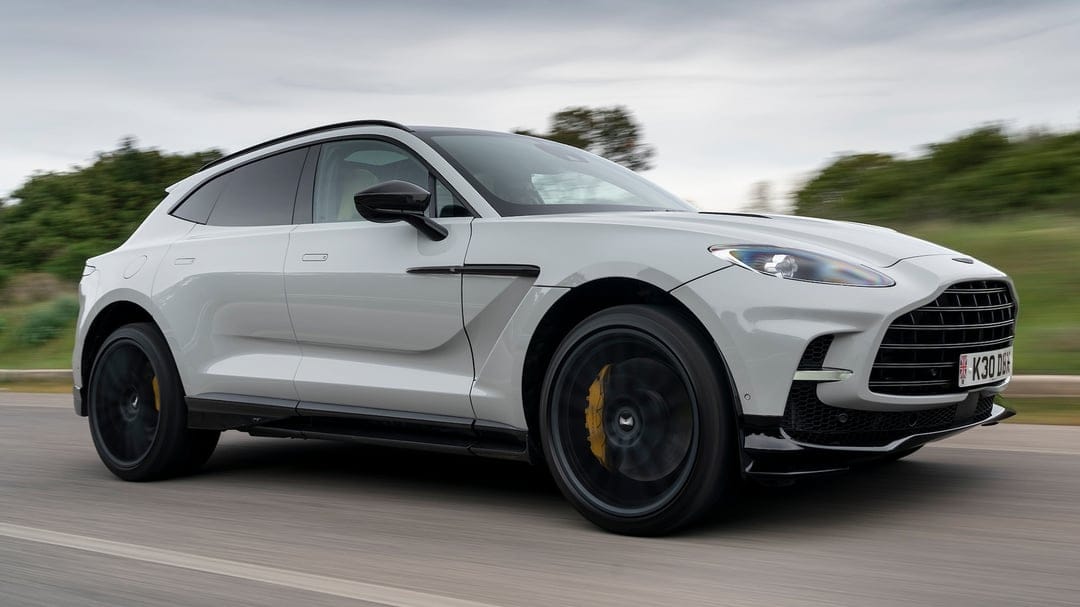 Silver Aston Martin DBX707 driving on a road with motion blur, black wheels, and yellow brake calipers.