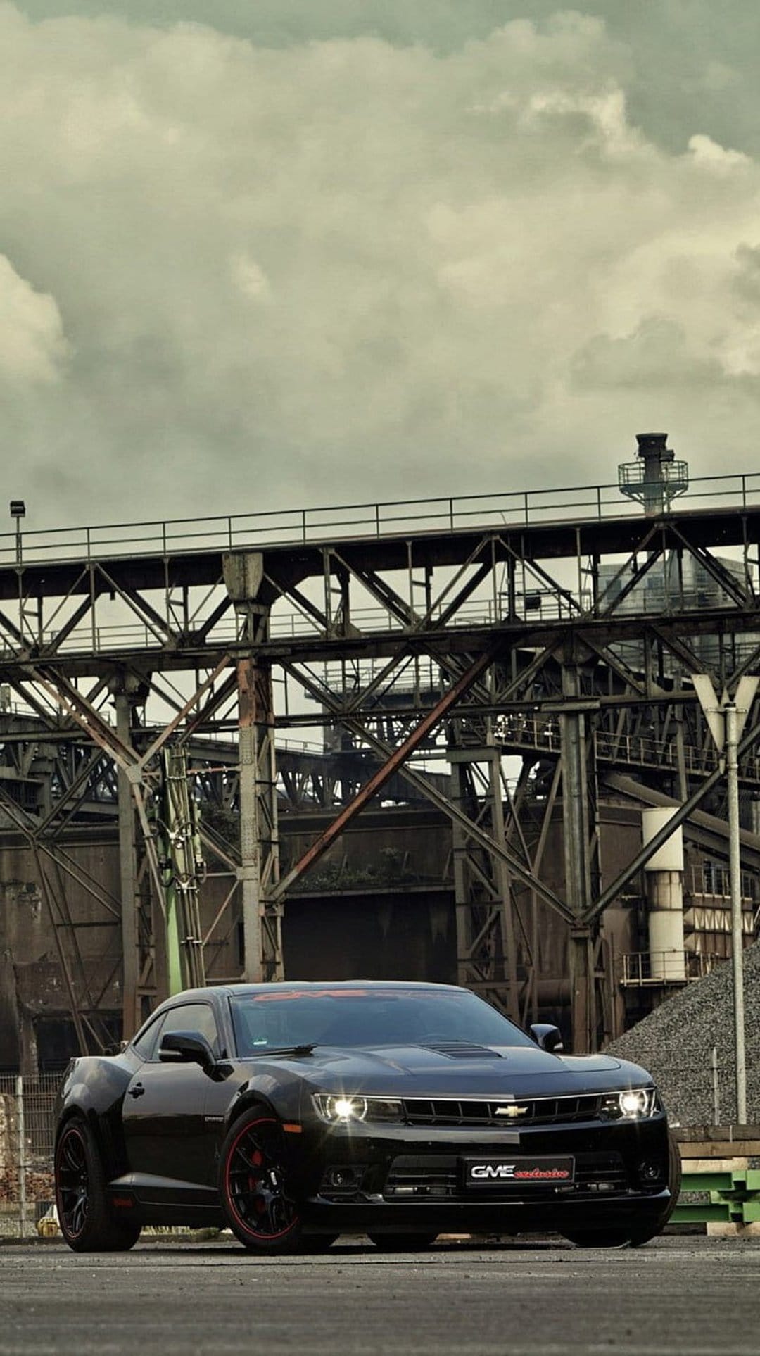Black Chevrolet Camaro parked near rusted factory pipes under a dark cloudy sky in a vertical composition.