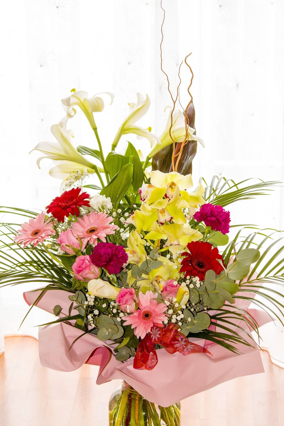 White lilies and yellow orchids mingle with red gerberas in a glass vase wrapped in pink paper and a red ribbon.