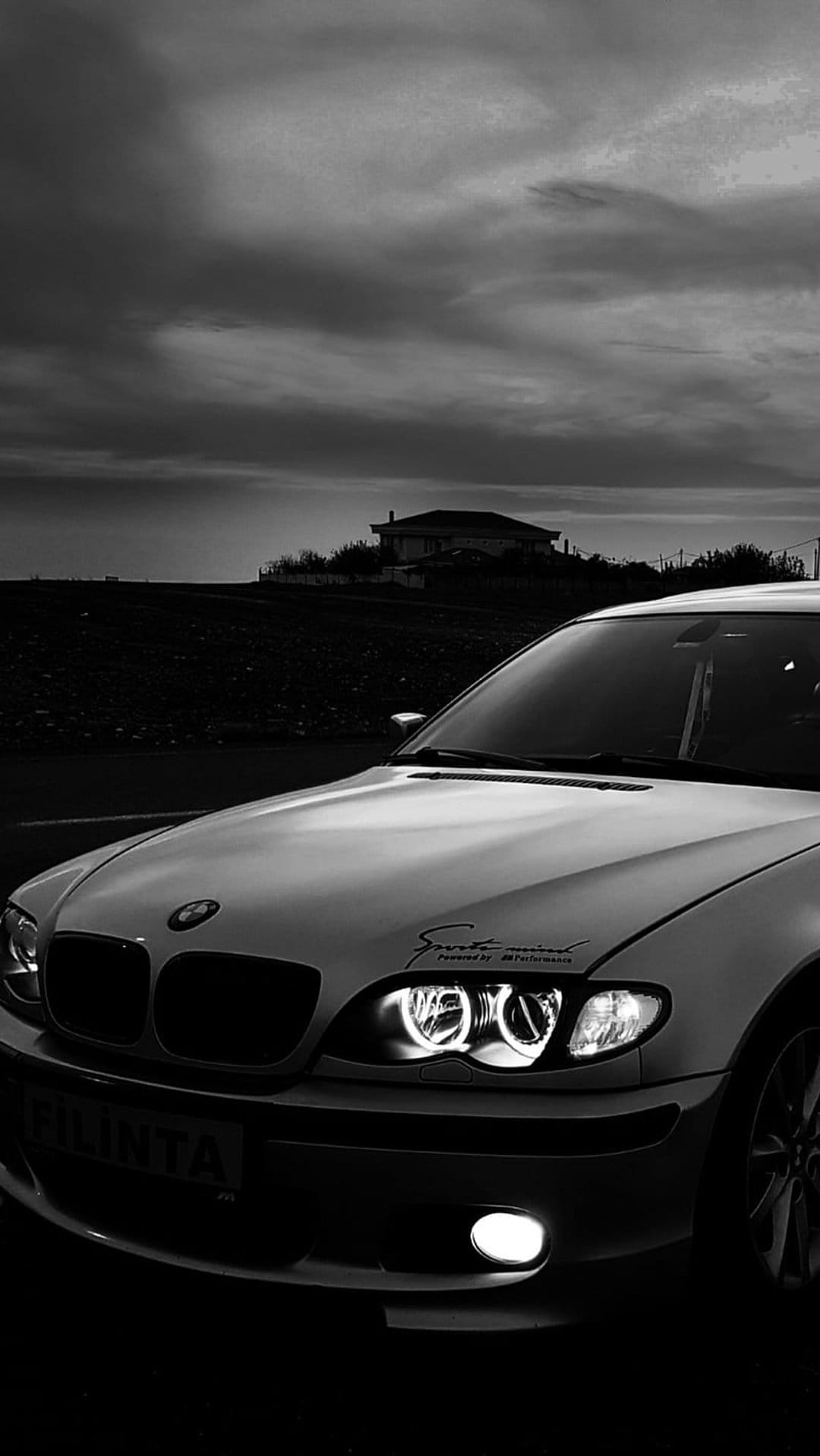 BMW E46 headlights shine from a low angle against a dark cloudy sky in a black and white vertical composition.