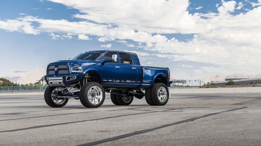 Blue lifted dually pickup truck with chrome wheels and suspension on a paved lot under a cloudy sky.