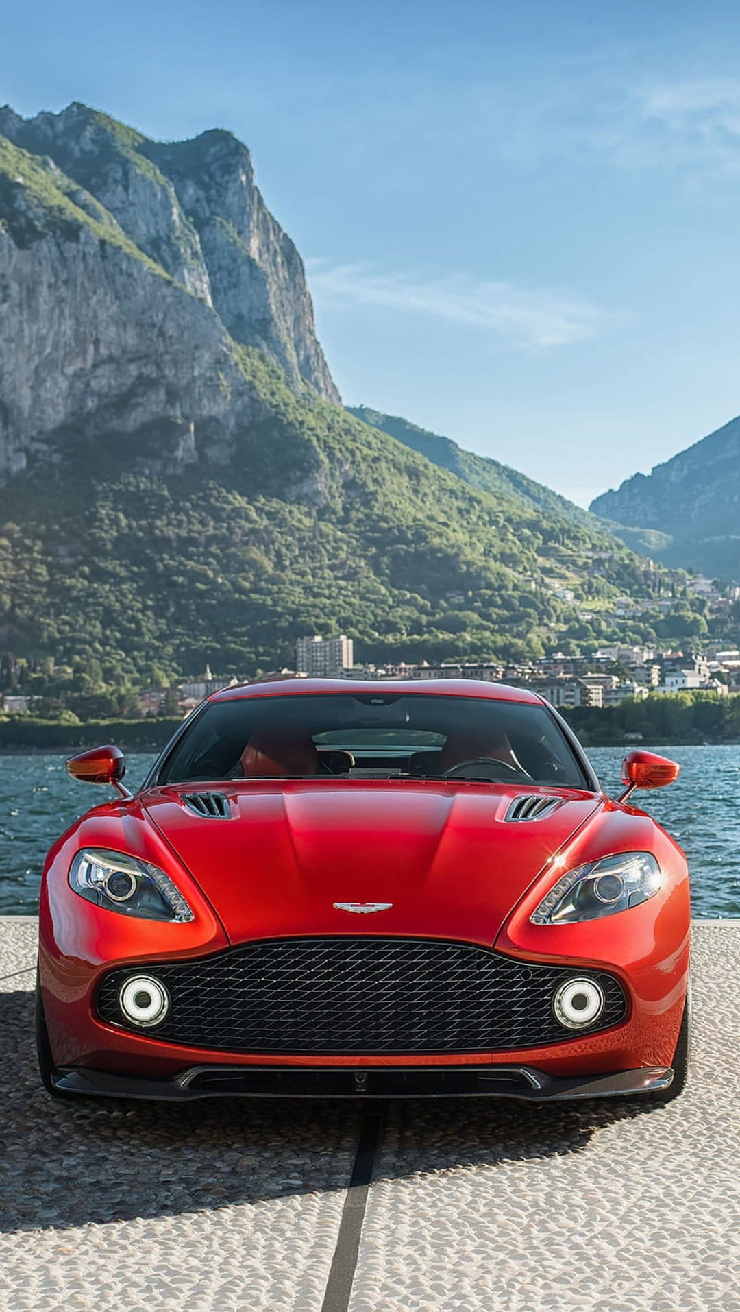 Red Aston Martin Vanquish parked on a stone quay with turquoise lake water and green mountain peaks behind it.