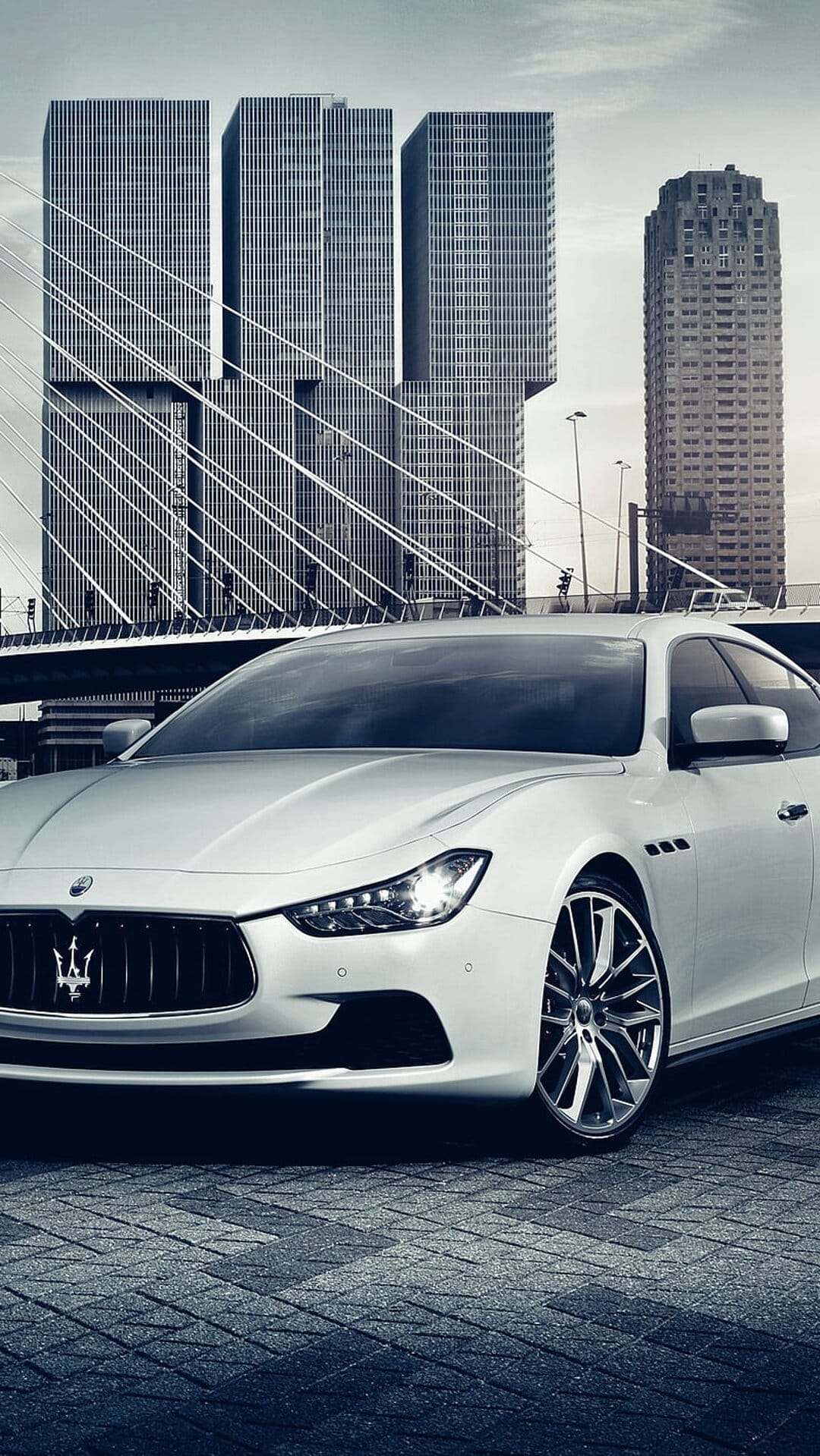 White Maserati Ghibli parked on cobblestones against a backdrop of tall glass skyscrapers in downtown Rotterdam.