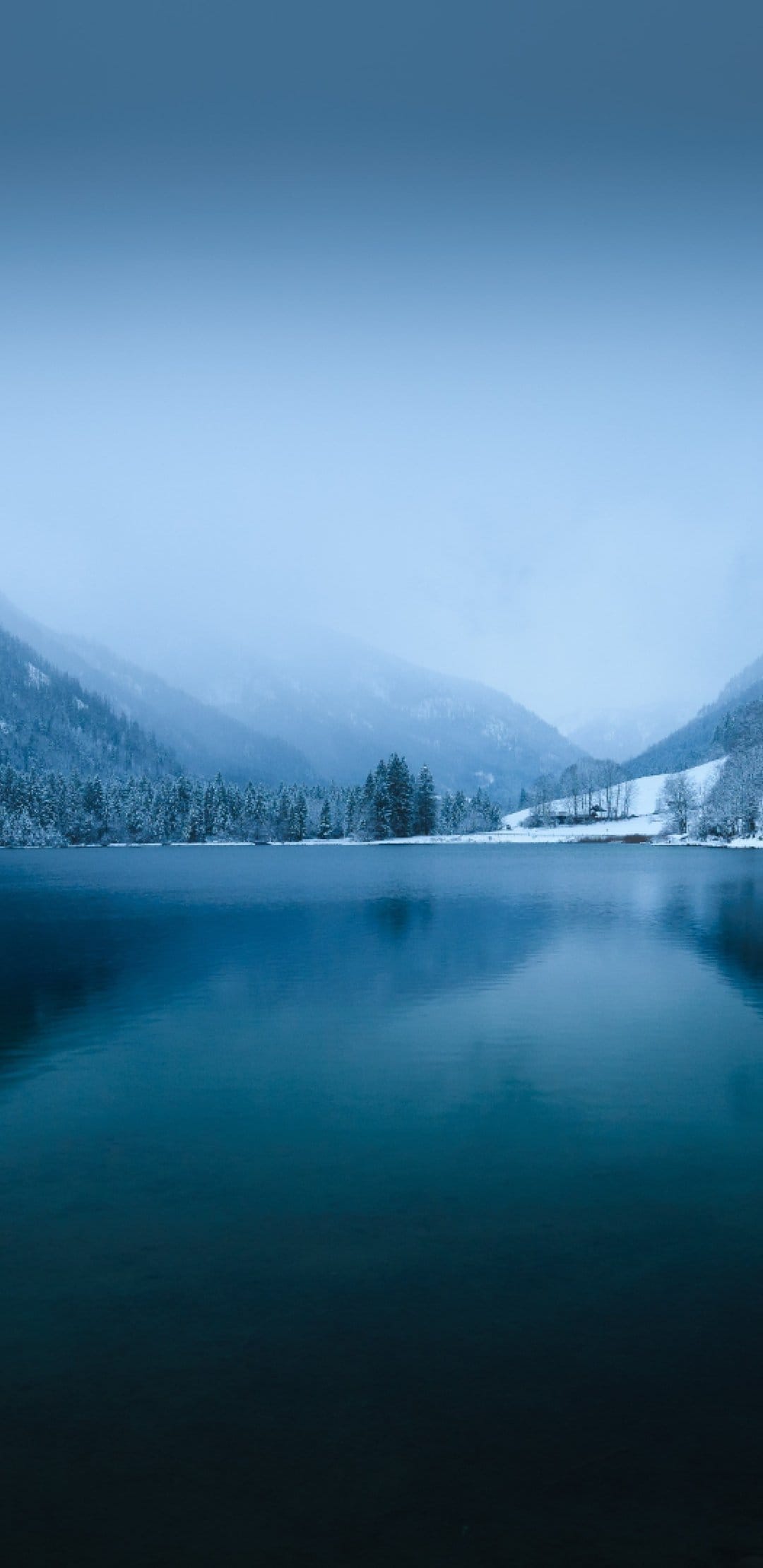 Blue mountain lake reflects snow-covered peaks and a misty evergreen forest under a soft, overcast winter sky.