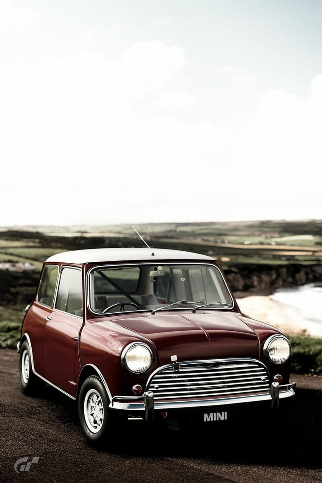 Maroon Mini Cooper with a white roof parked on a coastal dirt road near green hills under a bright cloudy sky.
