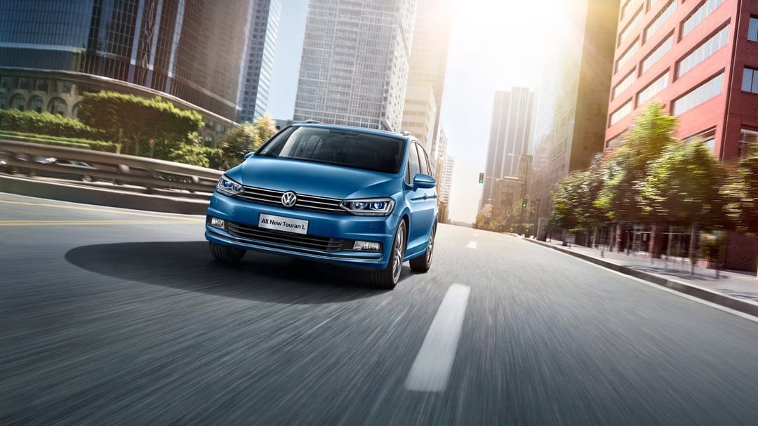 Blue Volkswagen Touran L driving on a city street with motion blur and bright sunlight hitting skyscrapers.