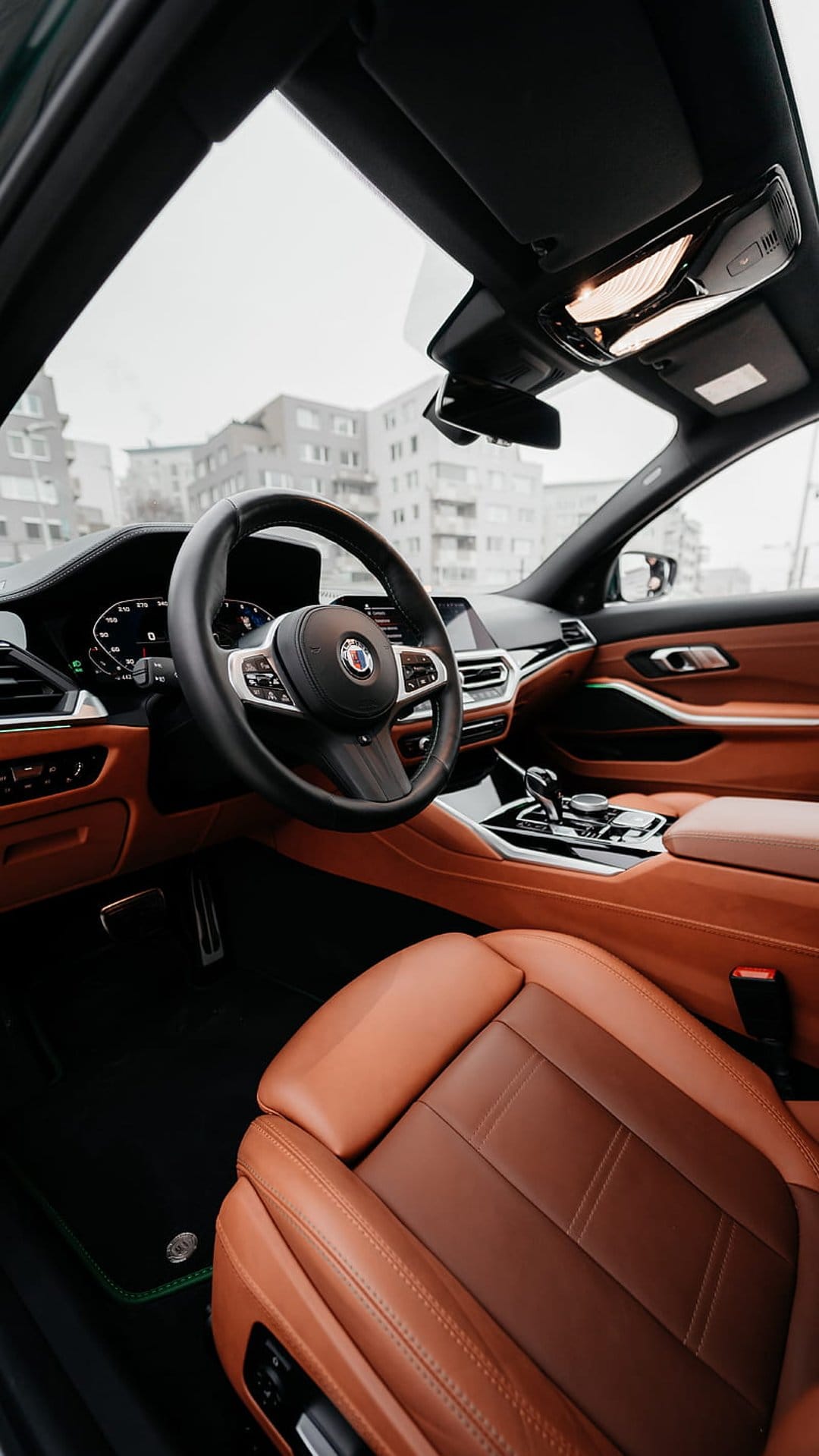 Brown leather car seats and a leather-wrapped steering wheel inside a luxury vehicle cabin with urban views.