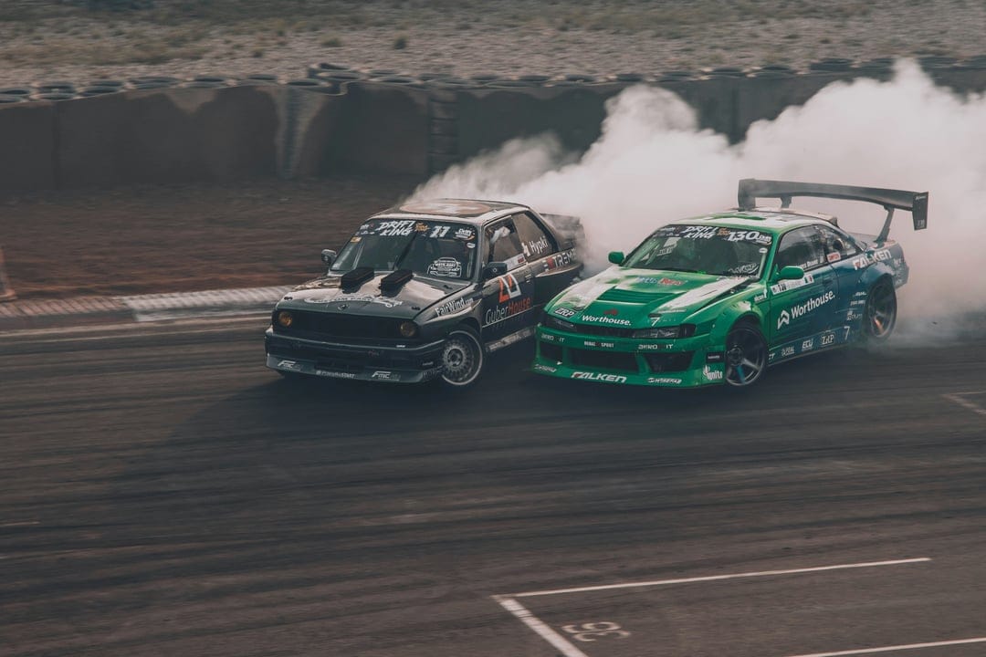 Black and green drift cars slide sideways on a track while emitting heavy white tire smoke in a vertical view.