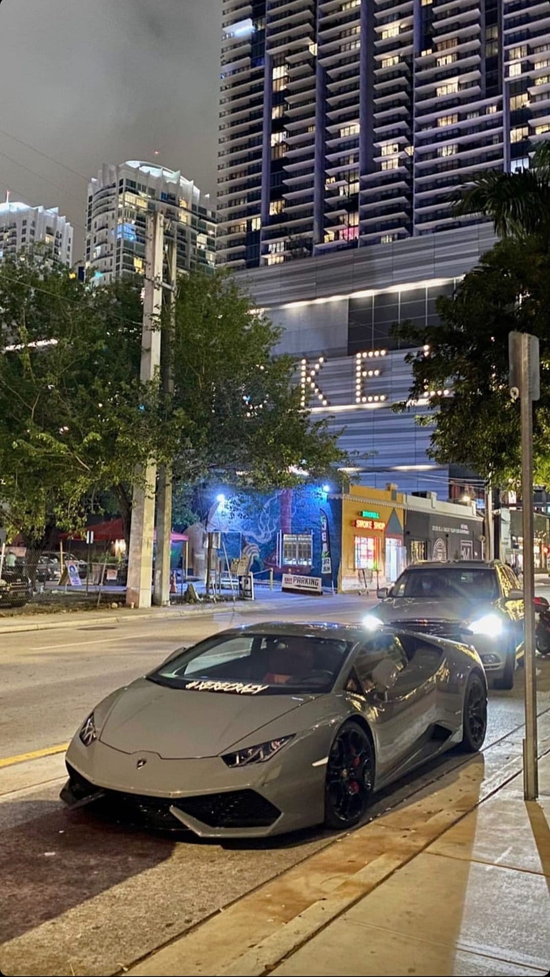 Grey Lamborghini Huracan parked curbside on a night city street with a glowing skyscraper in the background.