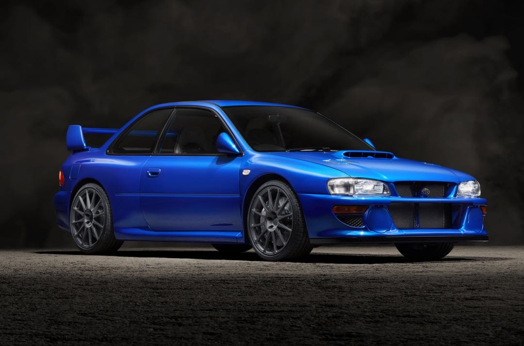 Blue Subaru Impreza STI coupe with charcoal wheels parked in a dark smoky studio with dramatic overhead lighting.
