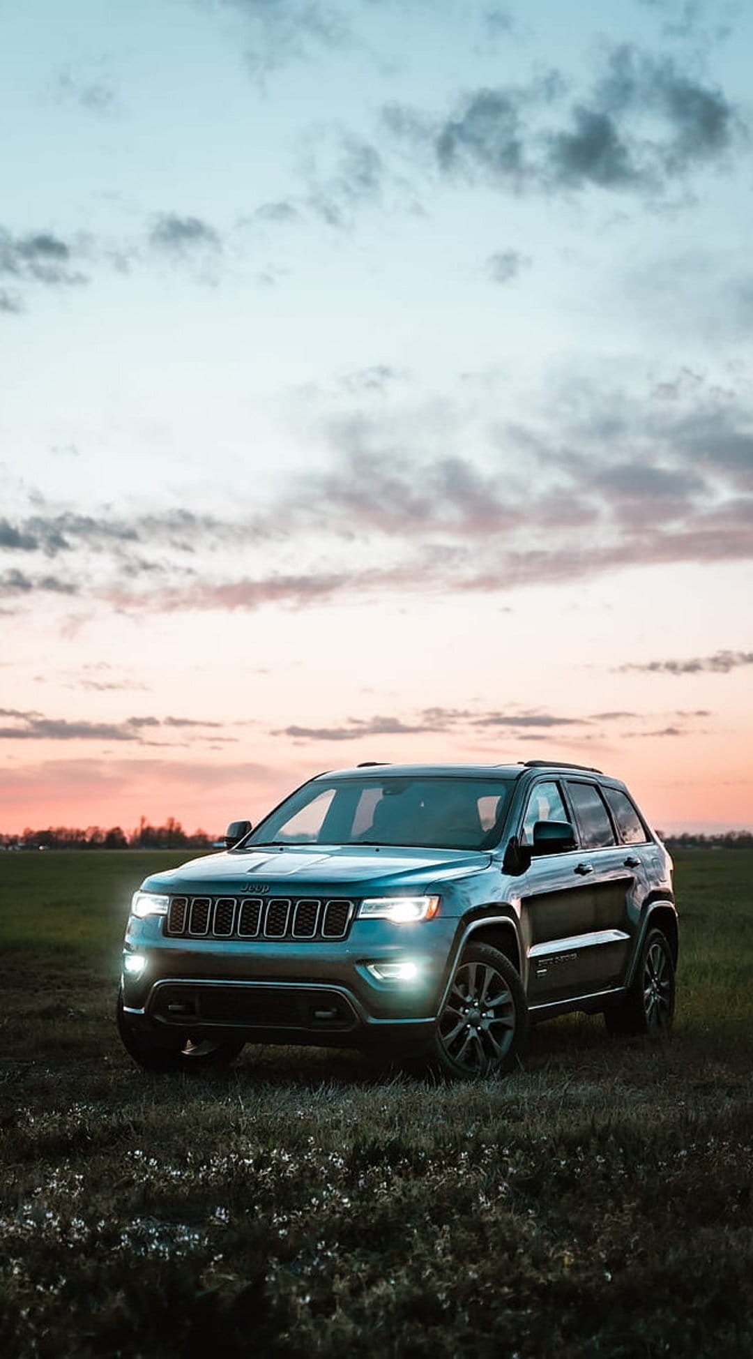 Jeep Grand Cherokee SUV parked in a grassy field with headlights on under a pink and orange sunset sky.