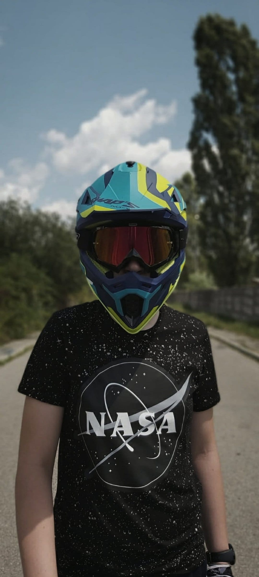 Person in a NASA t-shirt and racing helmet stands on an asphalt road with green trees and a blue sky behind them.
