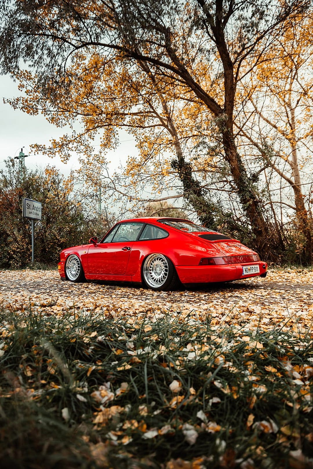 Red Porsche 911 classic car parked on a road covered in yellow autumn leaves surrounded by golden forest trees.