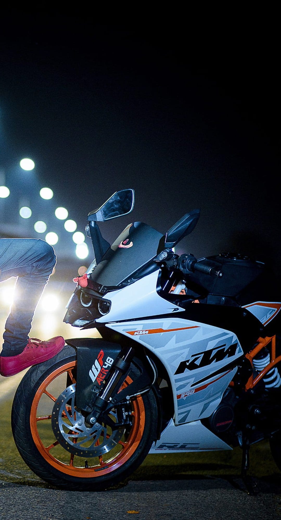 Red shoe rests on a KTM motorcycle tire with blurred golden city lights shining in the dark background.
