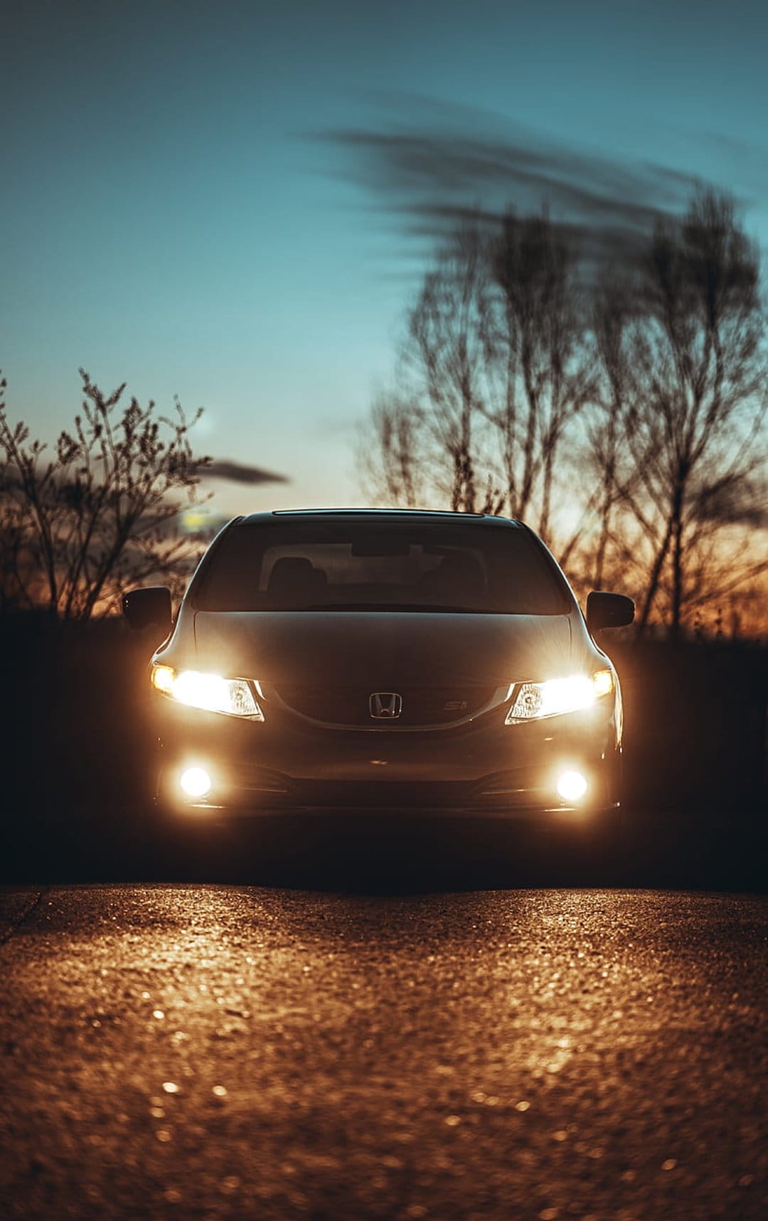 Honda Civic Si sedan with glowing headlights on a wet asphalt road under a cloudy evening sky with bare trees.