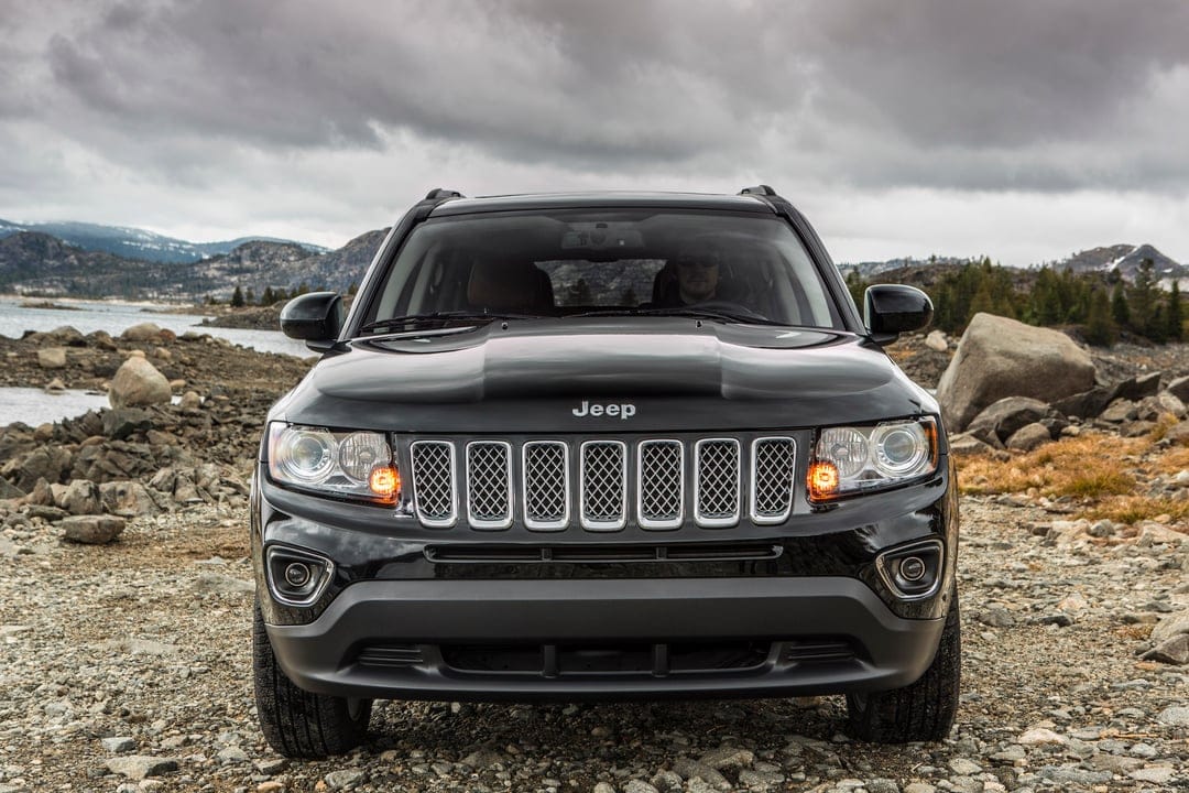 Front view of a black Jeep Compass on a rocky lakeshore with mountains and a cloudy grey sky behind it.