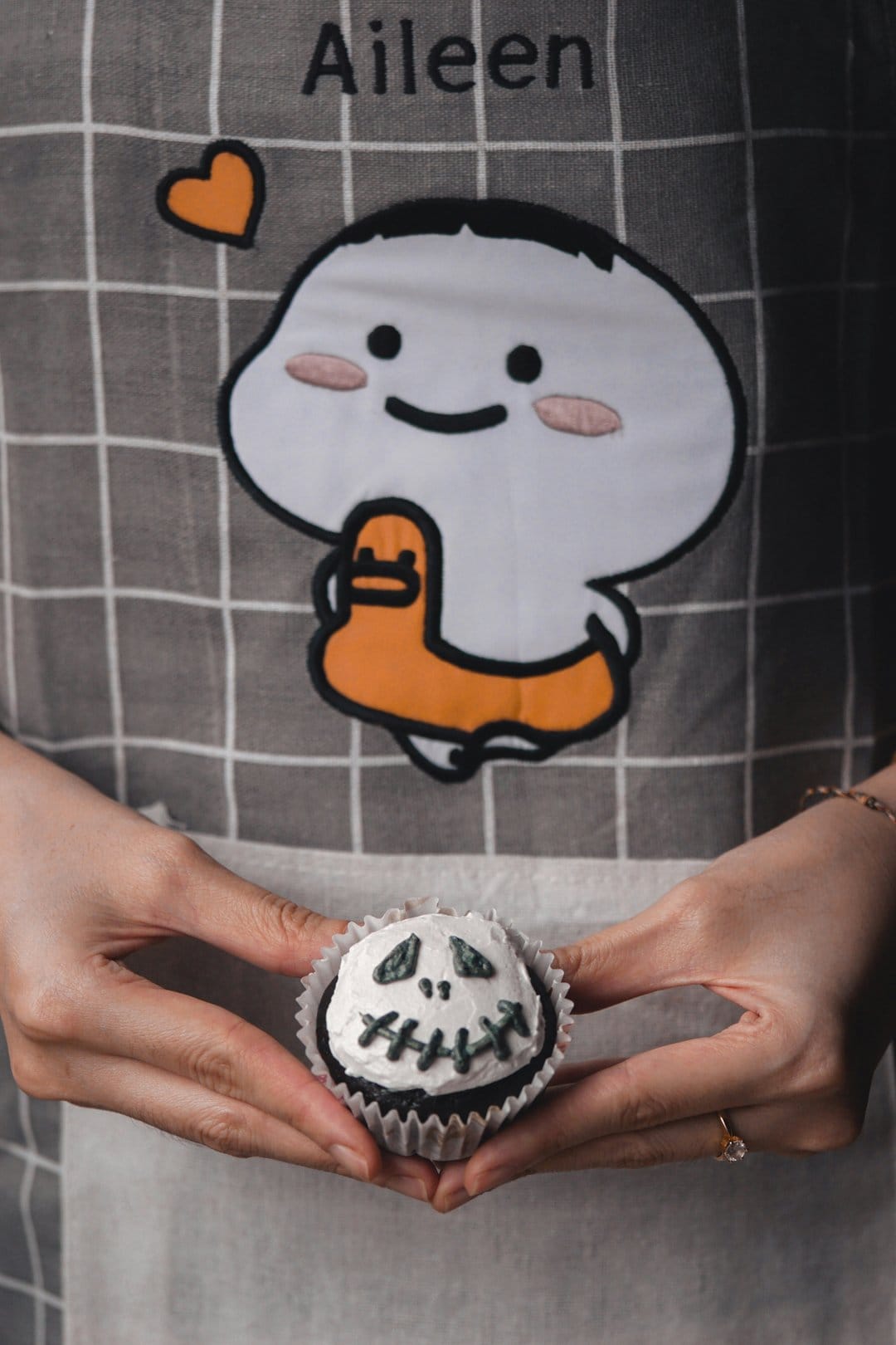 Jack Skellington cupcake held by a woman in a gray checkered apron with white and black frosting details.