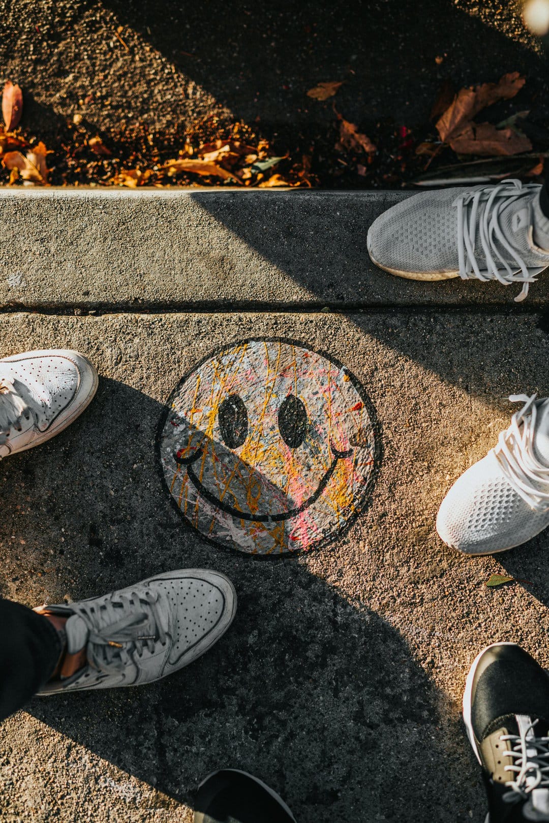 Smiley face drawing on a concrete sidewalk surrounded by diverse sneakers and scattered orange autumn leaves.