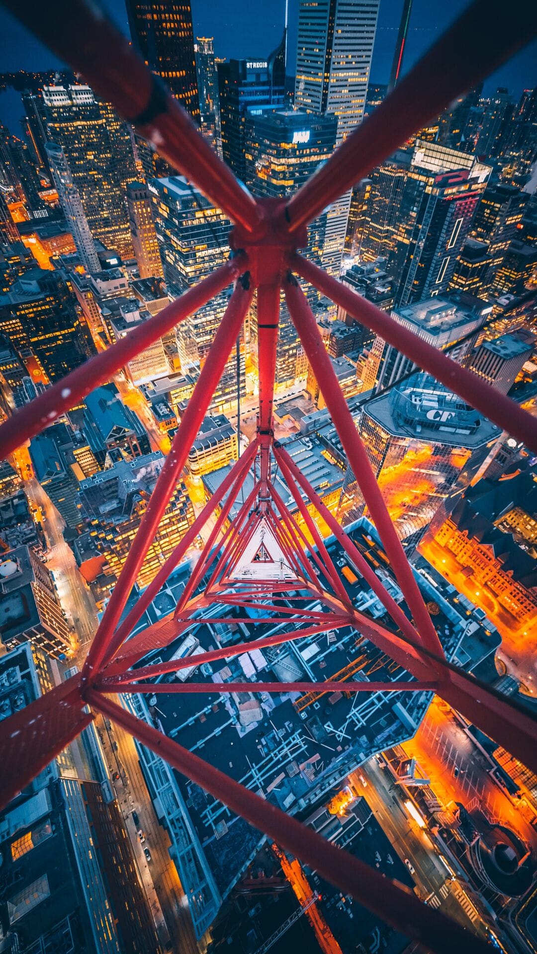 Red steel crane structure forms a geometric pattern above a lit city skyline during blue hour at night.