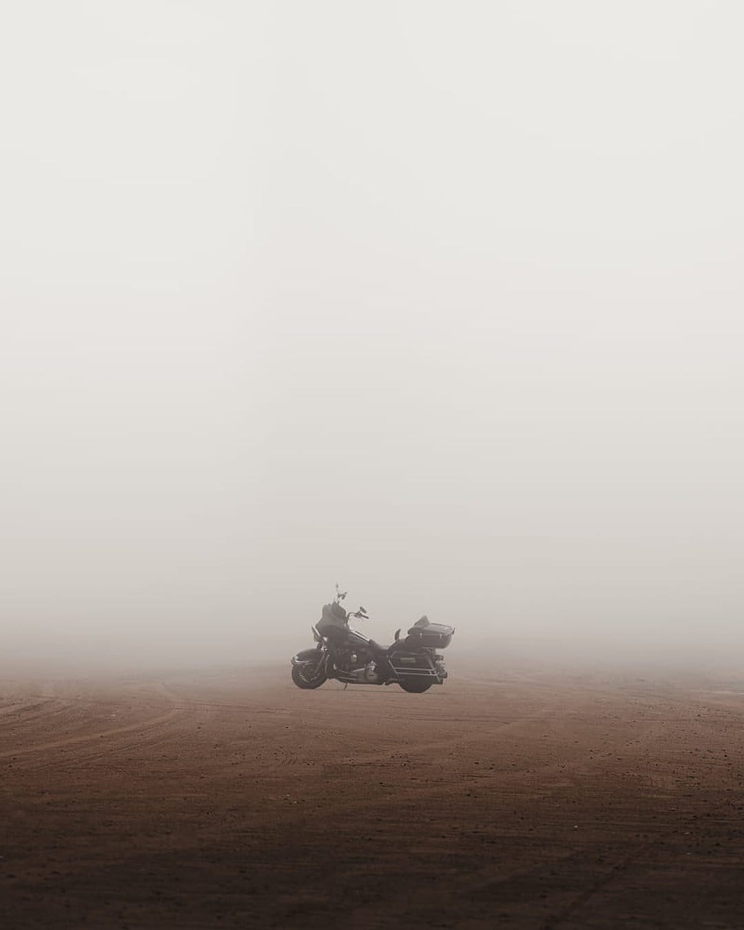 Vintage motorcycle parked on a dusty dirt path surrounded by thick white fog and a hazy grey horizon line.