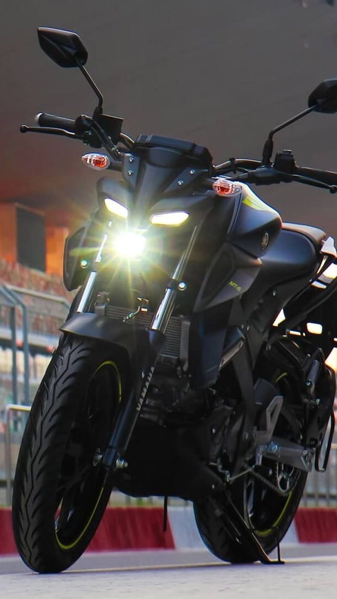 Yamaha MT-15 motorcycle front view with glowing LED headlights, black bodywork, and a blurred stadium background.