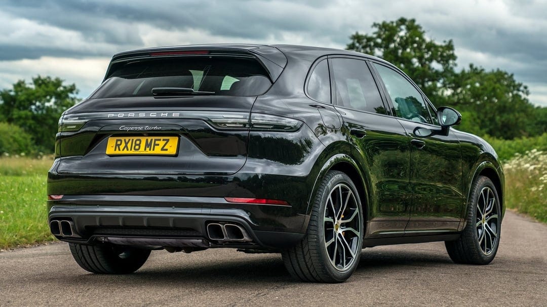 Black Porsche Cayenne Turbo SUV on a narrow road with lush green trees under a dramatic cloudy sky background.