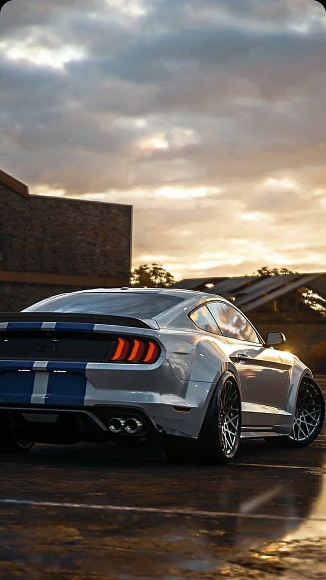 Silver Ford Mustang GT with blue stripes parked on wet pavement during a golden hour sunset with industrial clouds.
