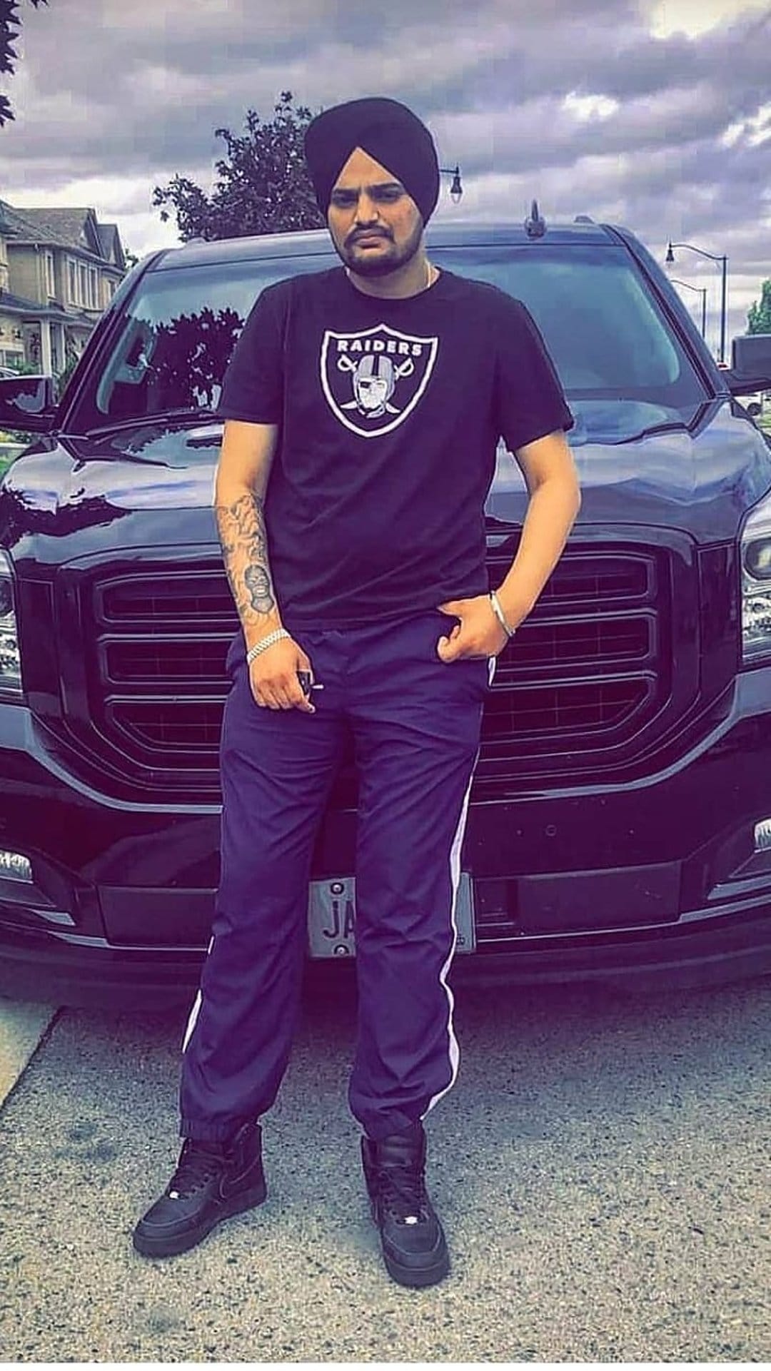 Sidhu Moose Wala stands by a black SUV wearing a black turban, Raiders t-shirt, and blue track pants outdoors.