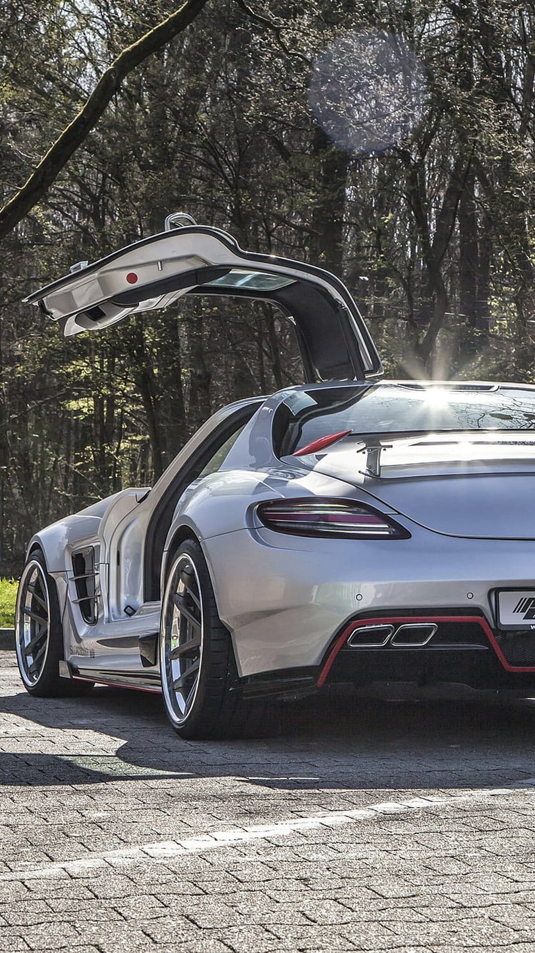 Silver Mercedes SLS AMG with open gullwing doors parked on a road within a sun-dappled forest landscape.