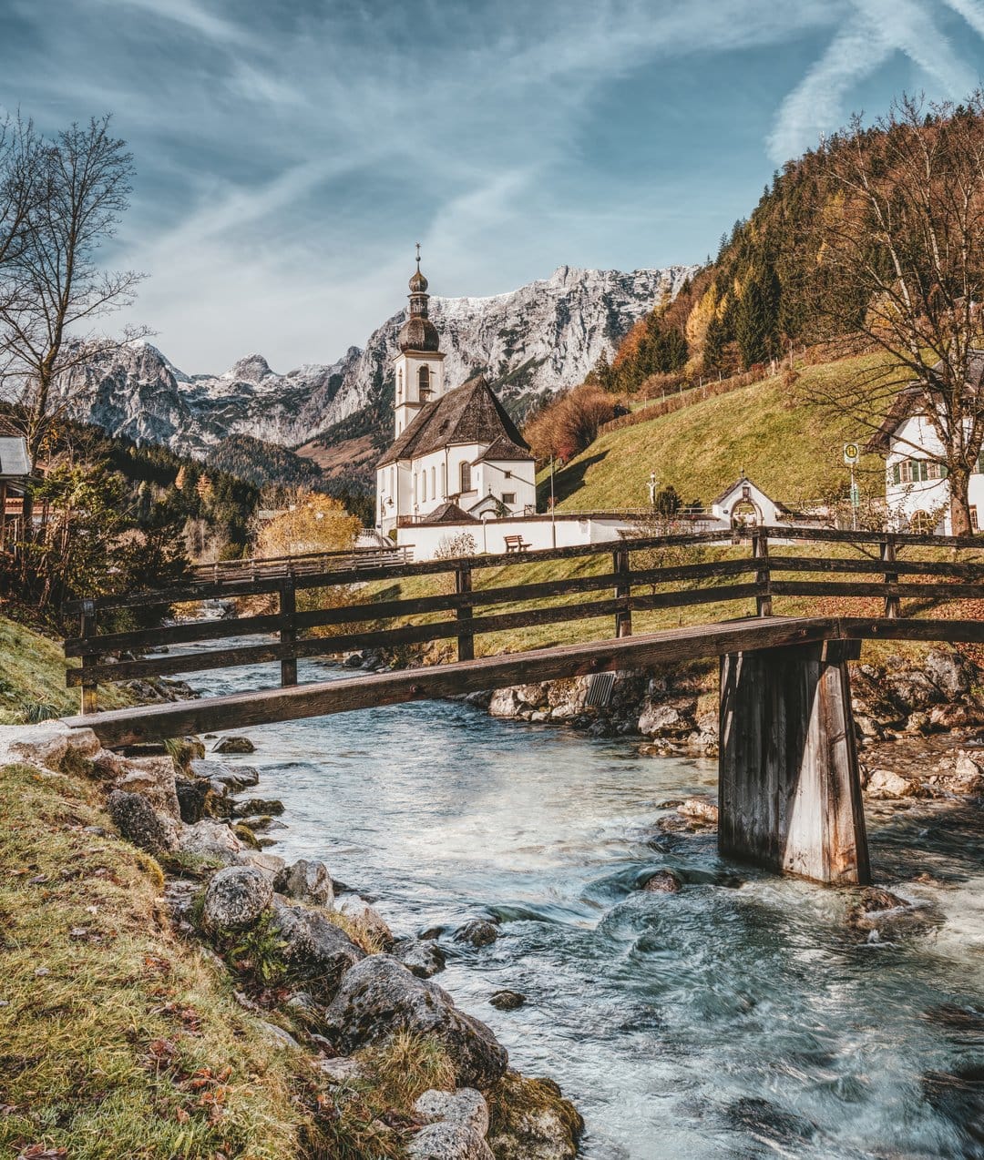 Alpine church and timber bridge overlook a rushing mountain stream surrounded by orange fall foliage and peaks.
