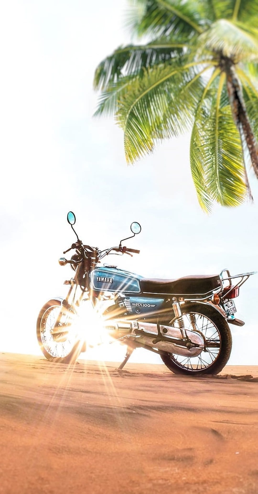 Blue Yamaha RX100 motorcycle parked on sun-drenched sand dunes with palm fronds and a soft lens flare.