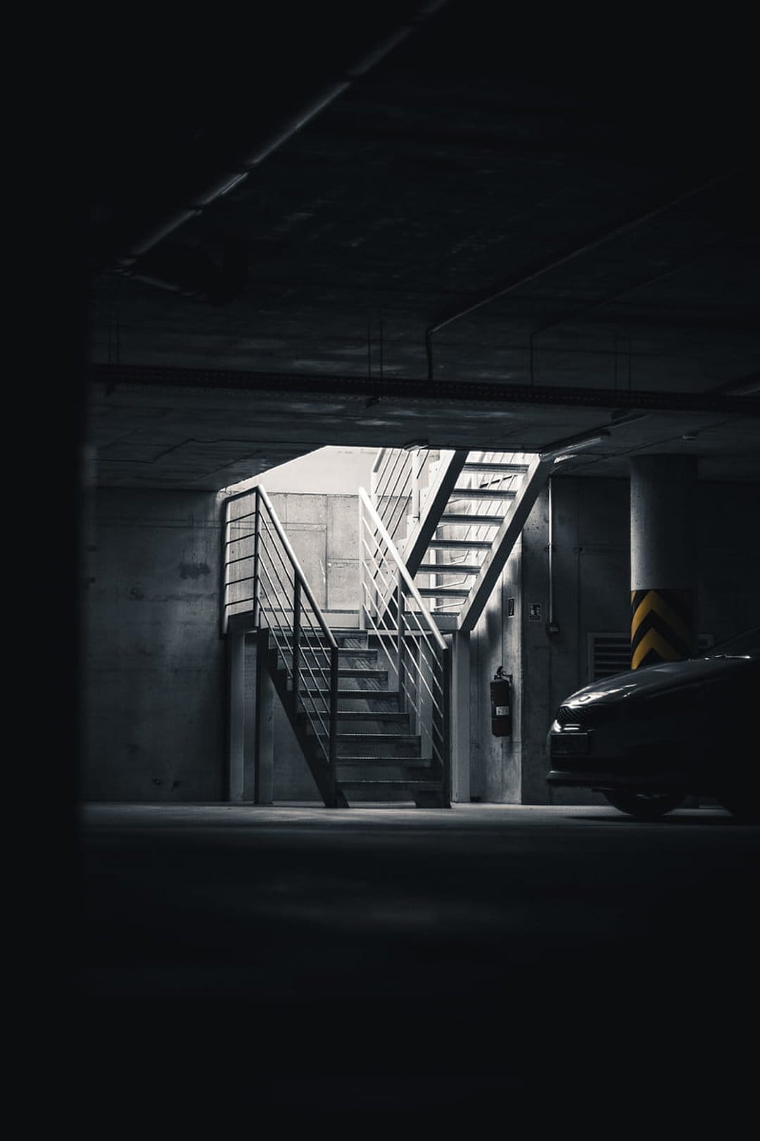 Metal stairwell in a dark concrete parking garage with a car partially visible under harsh overhead lighting.
