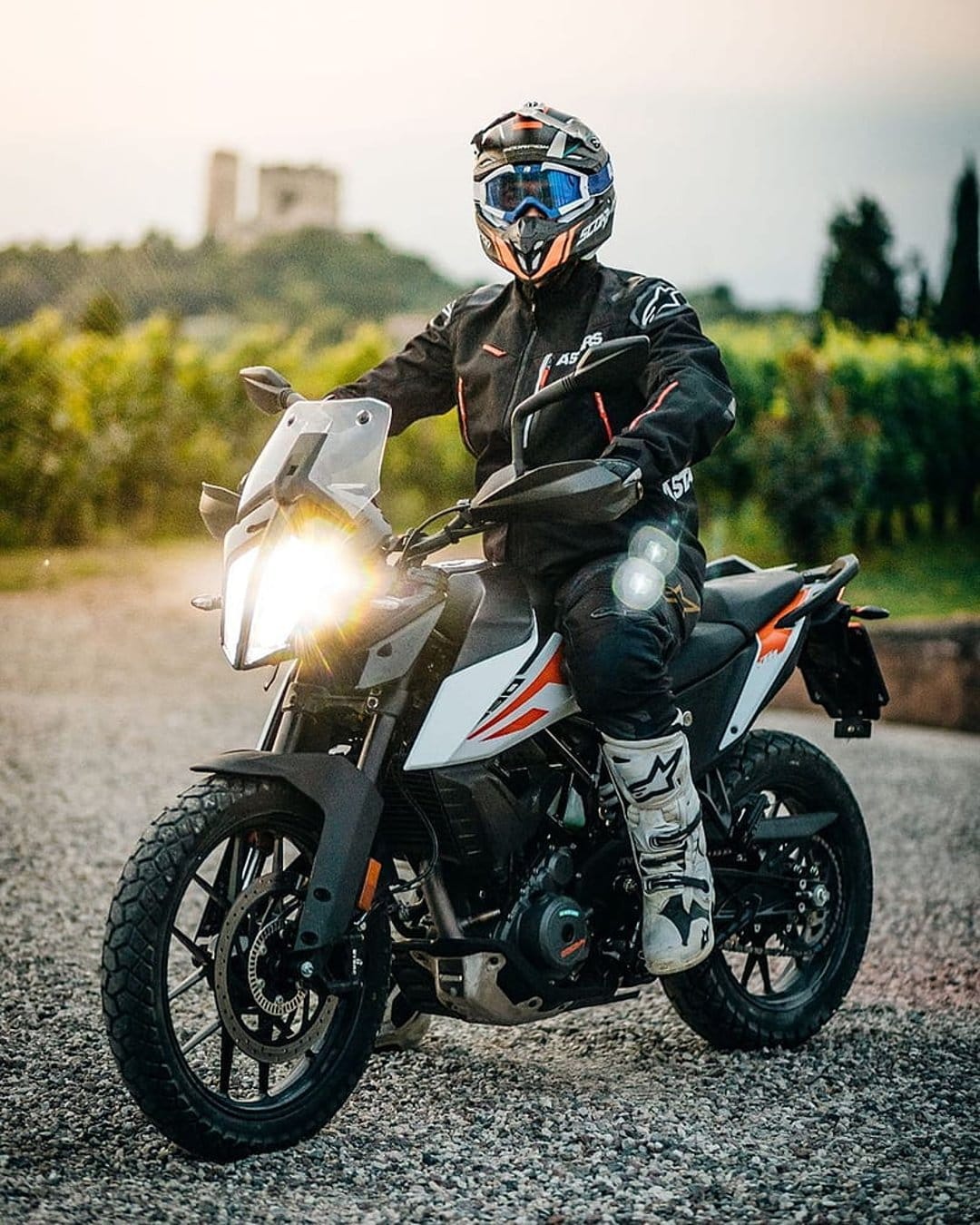 KTM adventure motorcycle rider sits on a gravel path at sunset with a glowing headlight and distant castle.