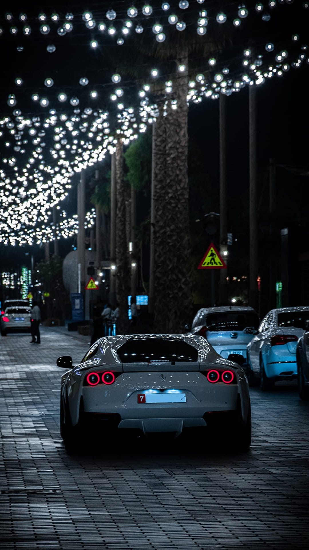 White luxury sports car with circular taillights parked on a street beneath strings of glowing festive lights.