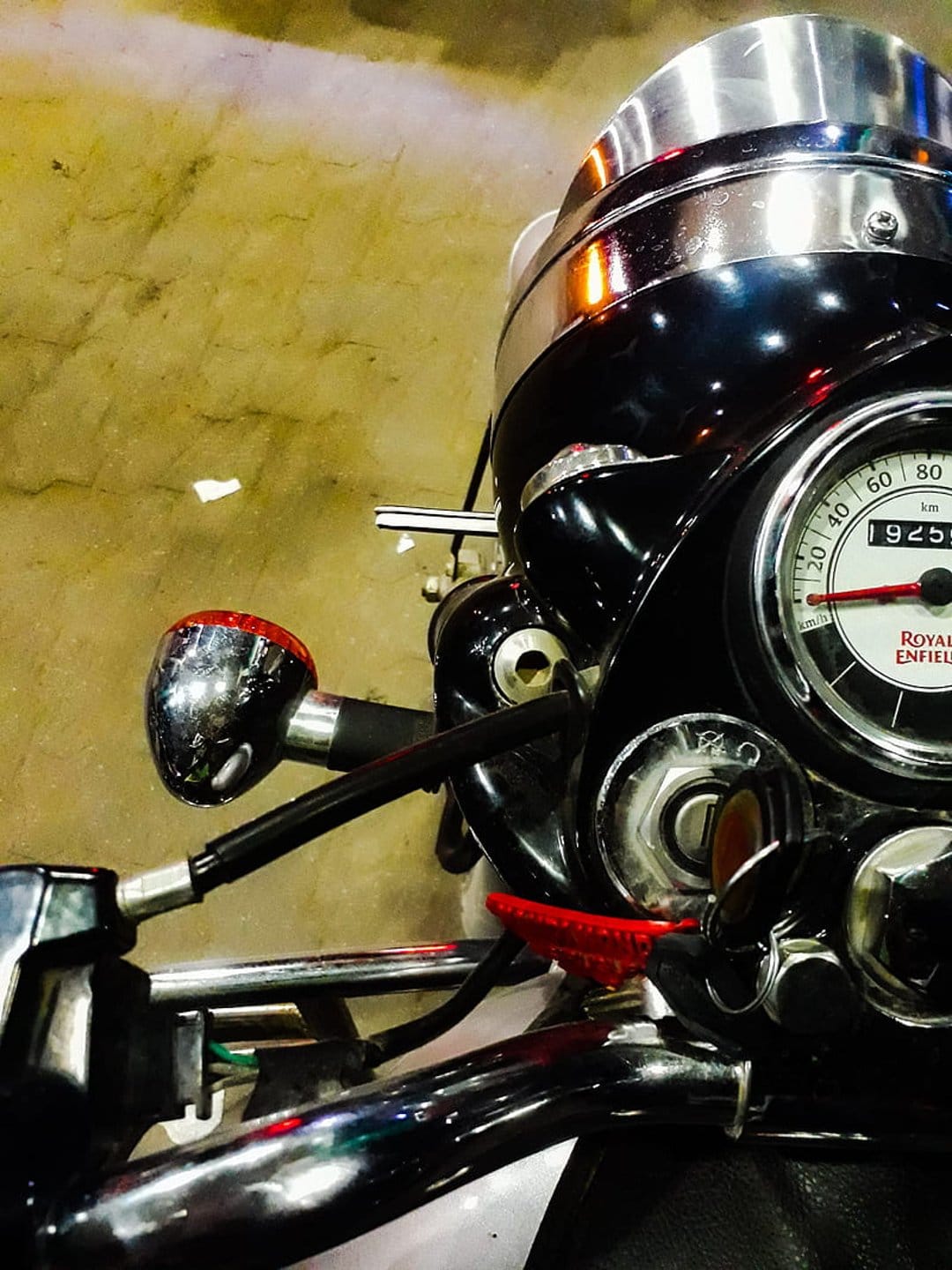 Royal Enfield chrome speedometer and handlebars against a textured wall with a red indicator light detail.