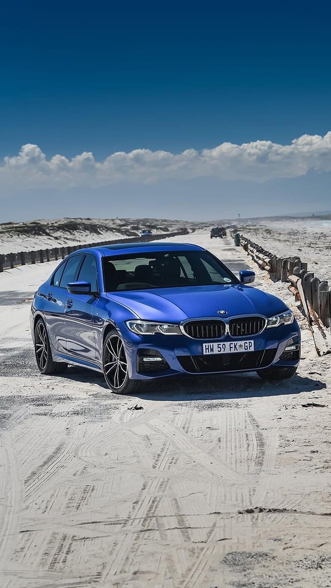 Blue BMW 3 Series sedan driving on a sandy beach road between wooden fences under a clear blue summer sky.